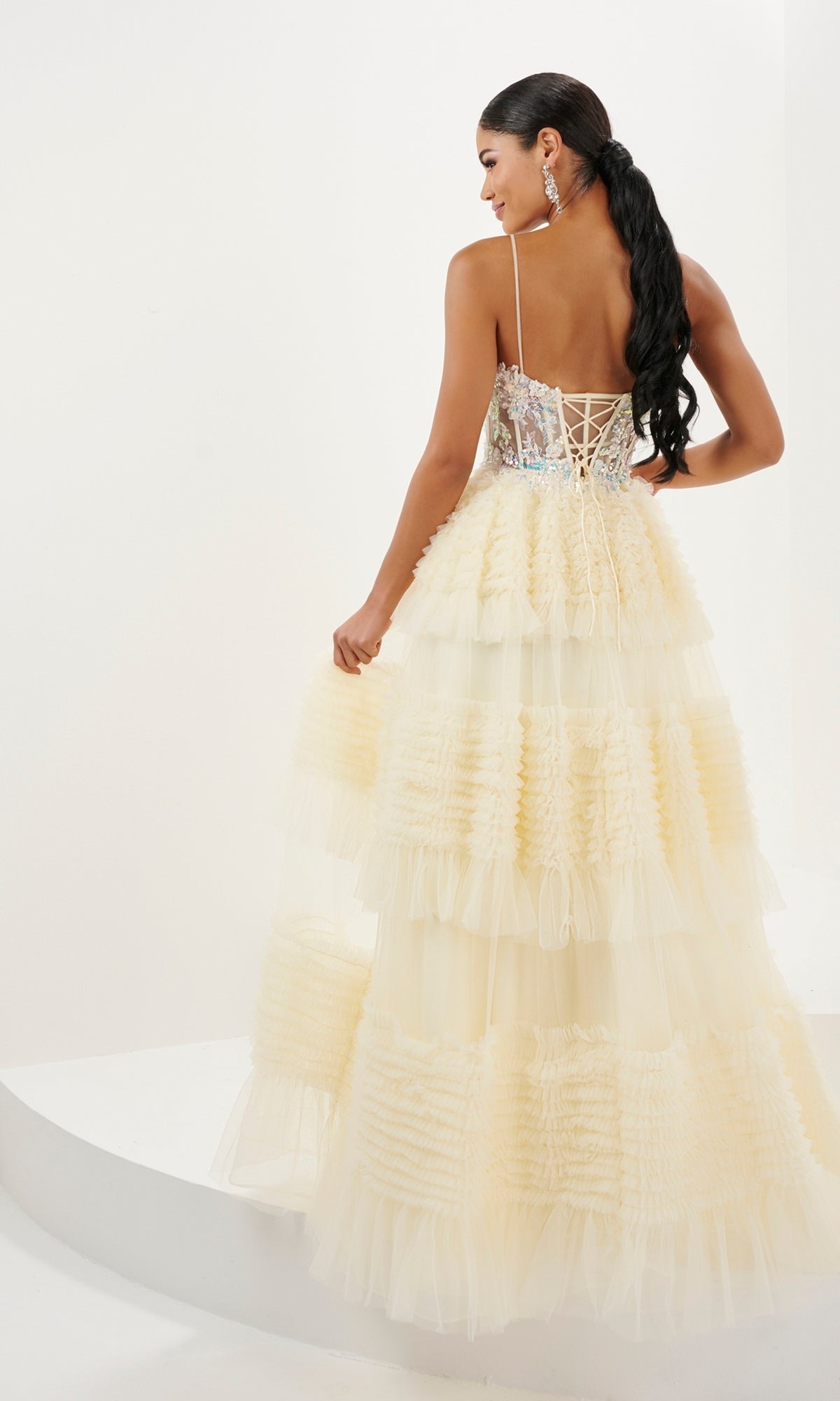 A woman in the Long Prom Dress 16054 by Tiffany, featuring an embellished v-neck, cream tiered ruffle skirt, a-line silhouette, and lace-up back, stands sideways holding the skirt.